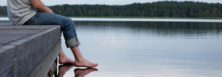 Chiropractic Muscatine IA Conditions A person sits on a wooden dock with their feet touching a calm lake, surrounded by a quiet forest under a cloudy sky.